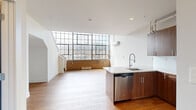 Located at 3715 Main Street in Philadelphia's vibrant Manayunk neighborhood, this two-story loft-style residence blends industrial character with modern finishes across two well-defined levels. The main floor opens into a bright, open-concept living and kitchen area bathed in natural light from a striking wall of large, grid-paned industrial windows that rise dramatically toward the ceiling. Original exposed brick accents the base of the window wall, adding warmth and historic texture to the space. Hardwood flooring runs throughout the main level, complementing the clean, contemporary aesthetic. The kitchen is anchored by a peninsula with a white quartz countertop and undermount sink, paired with rich walnut-toned cabinetry, stainless steel appliances—including a range, microwave, dishwasher, and refrigerator—and exposed ductwork overhead that nods to the building's industrial roots. A staircase leads to the upper level, where the generously sized primary bedroom features plush carpet, a sloped ceiling, a wall-mounted AC unit, and two closets. Two bathrooms serve the home, both finished with gray vanities, white countertops, and tile flooring, offering clean, functional design. This distinctive loft combines industrial heritage with thoughtful modern upgrades throughout.