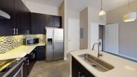 This well-appointed single-floor residence combines modern design with practical functionality throughout its thoughtfully laid-out spaces. The kitchen serves as a central highlight, showcasing dark espresso cabinetry that extends to the ceiling for generous storage, light quartz countertops, and a striking mosaic tile backsplash with a mix of neutral tones and subtle color accents. Stainless steel appliances, including a gas range and a French door refrigerator with a water and ice dispenser, complement the contemporary aesthetic. A kitchen island features a deep stainless steel sink with a modern gooseneck faucet, and a pendant light overhead adds a refined touch. Polished concrete floors extend through the kitchen and main living areas, providing a sleek, durable surface, while plush carpet adds comfort in the bedroom. The living area is bright and open, with a glass sliding door that leads to a private balcony with an outdoor view. The bathroom is clean and functional, offering a tiled soaking tub with a shower, a dark-cabineted vanity with quartz countertops, and direct access to a spacious walk-in closet equipped with wrap-around shelving and hanging rods. In-unit laundry is conveniently accessible, rounding out the practical amenities this residence has to offer.