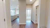 This single-floor residence is designed with a clean, contemporary aesthetic and a functional open-concept layout. The kitchen serves as a central focal point, featuring white shaker-style cabinetry extending to the ceiling, a granite or stone-topped island, stainless steel appliances including a refrigerator, range, microwave, and dishwasher, and pendant lighting that adds a refined touch. A subway tile backsplash and under-cabinet lighting further enhance the kitchen's polished appearance.

The living area flows seamlessly from the kitchen, offering generous open space bathed in natural light from multiple windows fitted with horizontal blinds. Wood-style plank flooring runs throughout the main living areas, providing a warm, cohesive look. A hallway with a structural column connects the main living space to the private areas of the unit.

Two bedrooms are each finished with plush carpeting, ceiling fans, and large windows that allow ample natural light. The bathroom features a white shaker vanity with stone countertops, a framed mirror with bar lighting above, and wood-style flooring. The neutral warm-beige wall palette throughout the unit creates a bright, welcoming ambiance well-suited for modern living.