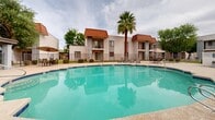 Located at 8605 N 59th Ave in Glendale, Arizona, this outdoor pool area serves as a central community amenity designed for relaxation and recreation. The centerpiece is a generously sized, irregularly shaped swimming pool with vivid turquoise water, framed by a broad concrete sundeck that provides ample space for lounging. Multiple lounge chairs and patio seating areas are positioned around the pool, offering residents a variety of spots to unwind under the Arizona sun. A covered pergola structure with a tile roof sits along one side of the deck, providing shaded seating beneath an open-air canopy — an ideal retreat from the heat. The surrounding buildings feature a Southwestern-influenced architectural style, characterized by stucco exteriors, red tile rooflines, and private balconies that overlook the pool area. Mature palm trees and deciduous greenery add natural texture and shade to the environment. The space is fully enclosed by a metal perimeter fence, ensuring privacy and security. A dedicated pool rules and safety signage station is centrally located near the deck. Overall, this pool courtyard presents a well-maintained, functional outdoor living space well-suited to the warm desert climate of the greater Phoenix area.