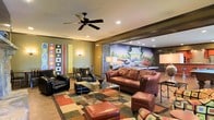 This single-floor lounge space exudes a lively, retro-inspired character through a carefully curated mix of bold design elements and comfortable furnishings. The main seating area anchors the room with rich brown leather sofas, armchairs, and ottomans arranged around a sleek glass-topped coffee table, all set atop a colorful geometric area rug that adds warmth and visual energy to the space. A ceiling fan with a dark finish overhead provides both function and a classic touch.

One of the most striking features is a large-scale vintage automotive mural that spans an adjacent wall, depicting classic cars in a nostalgic outdoor scene. This eye-catching artwork serves as a dramatic backdrop that ties the retro theme together seamlessly. Recessed lighting throughout ensures the space is well-lit, while wall sconces add a softer ambient glow.

An additional seating area is visible in the background, separated by an open partition, offering flexible use of the space for gatherings or quieter retreats. The walls feature bold, colorful abstract artwork, further reinforcing the dynamic and artistic personality of the interior. Warm-toned walls and hardwood-style flooring complement the overall design, creating an inviting and characterful environment that feels both social and relaxed.