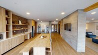 This single-floor communal space is thoughtfully designed to blend industrial and contemporary elements, creating a welcoming and functional environment. The layout is open and expansive, with light wood-tone flooring running throughout and recessed lighting providing consistent illumination across the space.

The kitchenette area is a central feature, equipped with stainless steel appliances including a full-size refrigerator and dishwasher, light wood cabinetry, open shelving, a sink, and a built-in cooktop. Dark brick walls serve as a striking backdrop, complementing the warm wood finishes and adding a distinctive industrial character to this zone. A large communal dining table provides ample space for group gatherings.

The lounge area offers a comfortable setting anchored by a wall-mounted electric fireplace framed in large-format tile. A natural wood plank accent wall adds warmth to this zone, which is furnished with plush seating and area rugs. Built-in open shelving provides display and storage functionality.

Large sliding glass doors and oversized windows flood the space with natural light while offering direct access to an outdoor patio area. The entry foyer features wainscoting-style wall paneling and decorative artwork, establishing a polished first impression. Overall, the space balances communal utility with a refined, modern aesthetic.