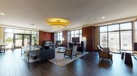 Located at 1910 Towne Centre Blvd in Annapolis, Maryland, this single-floor communal space offers a refined and welcoming environment defined by thoughtful design and high-end finishes. The main lounge area serves as the centerpiece, anchored by a striking gold drum pendant light that adds warmth and elegance to the room. Rich dark hardwood flooring runs throughout, complemented by deep mahogany wood-paneled walls featuring decorative circular motifs that lend a sophisticated, hotel-like aesthetic. Floor-to-ceiling windows line the perimeter of the lounge, flooding the space with abundant natural light and providing open views of the surrounding Towne Centre area. Seating is arranged in versatile groupings, including sofas, armchairs, and occasional tables, creating both intimate conversation areas and relaxed viewing zones centered around a wall-mounted flat-screen television. A built-in reception or service counter with warm granite countertops and rich wood cabinetry adds a functional yet polished focal point. An adjacent flex area features frosted glass sliding panel partitions with a sleek metal framework, offering a semi-private space suitable for meetings or events. Decorative touches such as framed artwork, lush indoor plants, and curated sculptural accents contribute to the space's upscale, inviting ambiance throughout.