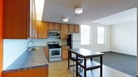 This single-floor space offers a thoughtfully designed open layout that seamlessly blends the kitchen and living areas. The kitchen is a standout feature, showcasing warm honey-toned wood cabinetry that extends to the upper walls, providing generous storage. Dark granite-style countertops run along the perimeter, complementing the full suite of stainless steel appliances, including a gas range, built-in microwave, and a full-size refrigerator. A deep basin sink is positioned beneath additional cabinetry, enhancing the kitchen's functionality. Elegant flush-mount crystal ceiling fixtures add a refined touch overhead. The space transitions naturally into an open dining area, where a dark wood high-top table and stools create an inviting casual dining setting. Large windows along the exterior wall flood the space with abundant natural light, offering views of the surrounding urban landscape. The neutral-toned walls and light concrete-style flooring provide a clean, modern backdrop throughout. The overall ambiance is modern yet welcoming, combining practical design elements with stylish finishes that make this a versatile and comfortable living space.