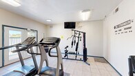 Located at 330 Waymore Dr in El Paso, TX, this fitness center is a single-room exercise space designed to support a well-rounded workout routine. The room is finished with light-colored tile flooring and cream-toned walls accented by teal baseboards, giving the space a clean and functional appearance. Overhead fluorescent and recessed lighting ensures the room is well-lit throughout.

The cardio area features two True-brand treadmills and a recumbent exercise bike, providing options for both high- and low-impact aerobic activity. A multi-station cable strength training machine, along with a Marcy weight bench, rounds out the equipment available for resistance training. A wall-mounted flat-screen TV is positioned for easy viewing during workouts. Motivational wall decals — including phrases like "No Pain No Gain" and "Be Stronger Than Your Excuses" — contribute to an encouraging atmosphere.

Natural light enters through a large set of double glass doors that open to an outdoor balcony, offering views of the surrounding El Paso landscape. A restroom is accessible directly from the exercise room, adding convenience for users. A fire extinguisher and safety equipment are also present. The space is compact yet efficiently organized to accommodate a variety of fitness activities.