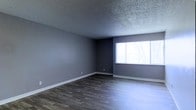 This single-floor space presents a straightforward and functional layout with a cohesive interior palette. The main living area is finished with dark hardwood-style plank flooring that runs throughout, providing a grounding contrast to the cool gray tones of the surrounding walls. The walls are painted in a uniform medium gray, lending the space a modern, neutral aesthetic that suits a variety of furnishing styles.

A large multi-pane window positioned along one wall serves as the primary source of natural light, brightening the interior and offering a view of the surrounding outdoor greenery. The natural light reflects off the flooring, enhancing the sense of openness within the room.

The ceiling features a textured popcorn finish, a common characteristic in residential construction, adding subtle visual depth overhead. Clean white baseboards run along the perimeter of the room, providing a crisp contrast to both the flooring and the wall color.

Overall, the space feels well-maintained and presents a blank canvas ready for personalization, with its neutral tones and durable finishes making it a practical and adaptable environment.