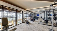 Located at 4020 Koehler Street in Houston's vibrant River Oaks/Washington Corridor neighborhood, this generously sized amenity clubroom is a standout social hub defined by dramatic architectural design and upscale finishes. The space is anchored by soaring double-height ceilings, floor-to-ceiling steel-framed windows that flood the interior with natural light, and direct views of the resort-style pool courtyard beyond.

Rich hardwood flooring transitions seamlessly into plush area rugs, delineating distinct zones throughout the open-plan layout. A central freestanding fireplace serves as a striking focal point, while exposed wood beam ceilings and an eye-catching geometric lattice ceiling installation add layers of texture and visual interest.

The lounge area features plush rust-orange swivel chairs and teal sofas arranged around a glass coffee table, complemented by a built-in upholstered window bench with accent pillows. A dedicated game zone includes a foosball table, shuffleboard, and billiards table beneath designer pendant and chandelier lighting. A bar area with counter seating and a media wall with a large flat-screen TV round out the entertainment offerings.

The space is further elevated by curated artwork, sculptural light fixtures, and a cohesive contemporary-meets-warm-industrial aesthetic that makes it equally suited for casual socializing or community gatherings.