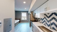 Located at 3408 S Budlong Ave in Los Angeles, this efficiently designed one-bedroom, one-bathroom unit offers a well-planned layout that maximizes every square foot. Light wood-look flooring runs throughout, complementing the contemporary interior palette of cool grays and crisp whites.

The bedroom area features a built-in bed frame with under-storage drawers, a dedicated desk and chair, and a large window with a roller shade that provides views of the surrounding Los Angeles neighborhood and fills the room with natural light. A wood-toned wardrobe adds practical storage.

The kitchen is thoughtfully outfitted with stainless steel appliances including a full-size refrigerator, electric range, and over-the-range microwave. A bold blue-and-white chevron tile backsplash adds visual interest, while a combination of white upper cabinets and wood-finish lower cabinetry with a built-in pantry tower provides ample storage. A white quartz-style countertop and integrated sink complete the space.

The bathroom showcases a walk-in shower with striking blue herringbone tile, white subway tile accents, a chevron-patterned floor, a wood-finish vanity, and brushed nickel fixtures, delivering a cohesive, design-forward finish throughout.