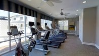 This single-floor fitness center is thoughtfully designed to provide a comfortable and functional workout environment. The space is dominated by a row of high-quality cardio machines, including treadmills and stationary bikes, each equipped with individual monitors for an engaging exercise experience. Large floor-to-ceiling windows line one side of the room, flooding the interior with abundant natural light and offering views of the surrounding outdoor area, creating an open and energizing atmosphere. Two ceiling fans are installed overhead, ensuring consistent airflow and comfort throughout workout sessions. The neutral-toned walls are accented with a piece of abstract artwork, adding a touch of character to the space. Additional windows fitted with slatted blinds on the adjacent wall allow for further light control and ventilation. The flooring features a durable, low-pile carpet suited for fitness use, and the overall layout is spacious enough to accommodate multiple users simultaneously. Recessed lighting supplements the natural light for evening or low-light use. The fitness center presents a clean, modern aesthetic that balances practicality with a welcoming ambiance.