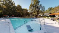 Located at 3190 Skinner Mill Rd in Augusta, Georgia, this outdoor amenity area centers around a generously sized swimming pool designed for both relaxation and recreation. The pool features built-in water jets, submerged lounge chairs, and multiple entry points via stainless steel handrails, making it accessible and inviting for a wide range of users.

Surrounding the pool is a broad, light-colored concrete deck that provides ample space for lounging and socializing. A row of cushioned chaise lounges lines one side of the pool, positioned to take full advantage of natural sunlight throughout the day. On the opposite end, an outdoor dining area offers a table and chairs shaded by a vibrant orange umbrella, ideal for casual gatherings.

The adjacent clubhouse or amenity building is a single-story structure with a pitched roof, white exterior, and columned entrance, complemented by manicured landscaping including ornamental trees and neatly trimmed shrubs. A wide concrete pathway connects the building to the pool deck, flanked by a black metal fence that defines the perimeter of the space.

The entire area is embraced by a canopy of mature, towering trees that provide a natural backdrop and contribute to the tranquil, park-like atmosphere. The combination of well-maintained grounds, functional amenities, and abundant greenery creates a welcoming outdoor retreat.