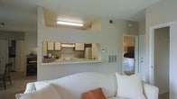This single-floor residence is designed with an efficient, open layout that maximizes the use of available space. The living area serves as the central hub of the home, furnished with a comfortable white sofa accented by warm throw pillows, creating an inviting atmosphere. A defining architectural feature is the pass-through window connecting the living area to the kitchen, which adds visual openness and allows for easy interaction between the two spaces. The kitchen is equipped with standard appliances, including a white electric range and a refrigerator, complemented by wooden upper cabinetry and a white countertop that extends to form a convenient serving ledge along the pass-through. An overhead fluorescent fixture provides functional task lighting above the kitchen area. The neutral, light beige wall tones throughout contribute to a cohesive and airy feel. A doorway visible from the living area leads to the bedroom, which is furnished with a bed and offers a carpeted floor, maintaining the home's understated, practical aesthetic. The layout promotes a natural flow between living, cooking, and sleeping spaces.