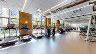 This fitness center offers a thoughtfully designed, multi-zone layout that accommodates a wide range of workout activities. The main cardio area is a standout feature, characterized by floor-to-ceiling windows framed by warm natural wood accent walls, flooding the space with abundant natural light and offering views of the surrounding greenery and cityscape. The open floor plan comfortably houses a full row of treadmills, elliptical trainers, stair climbers, and stationary cycling bikes, all arranged to maximize visibility and airflow. Large wall mirrors along one side visually expand the already generous space.

An adjacent strength training area provides additional workout versatility, equipped with a rowing machine, cable machine, weight bench, and free weights. Glass partition walls connect this zone to the broader facility while maintaining a sense of openness. A dedicated studio room features resilient flooring, wall-mounted ballet barres, full-length mirrors framed in gold accents, and large windows overlooking an outdoor courtyard. This multi-purpose studio is well suited for group classes, stretching, or individual training sessions. Linear LED lighting runs throughout the facility, providing consistent, bright illumination across all areas. The overall design balances modern aesthetics with functional fitness infrastructure.