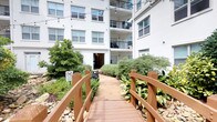 This single-floor residence is situated within a multi-story building and offers a thoughtfully designed living experience both inside and out. The exterior courtyard is a standout feature, showcasing a wooden footbridge flanked by river rock beds, mature shrubs, and tropical foliage. String lights are strung overhead, creating an inviting atmosphere, while upper-level balconies with black iron railings overlook the landscaped grounds.

Inside, the unit opens to a well-designed layout with dark hardwood-style flooring that flows through the main living areas, contrasting cleanly with the light-toned walls and white trim. The kitchen is equipped with stainless steel appliances, including a range, microwave, dishwasher, and refrigerator, complemented by white cabinetry, a gray subway tile backsplash, and a light-colored countertop peninsula that separates the kitchen from the living area. The open sightlines between the kitchen and living room enhance the sense of space and connectivity.

The living area benefits from natural light through large windows and is finished with a ceiling fan. A hallway connects the main living spaces to additional rooms, including a bedroom visible from the corridor. Storage is well-addressed with louvered and panel-style doors providing access to closets and utility areas throughout the unit.