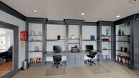 This single-floor business center offers a well-appointed workspace designed with both functionality and style in mind. The centerpiece of the room is a expansive built-in workstation finished in a sophisticated gray tone, featuring two computer stations, integrated shelving units, and ample cabinet storage below. The open shelving displays books and decorative accents, lending the space a professional yet inviting character. Bold dark crown molding lines the ceiling throughout, creating a strong architectural contrast against the light gray walls and adding a sense of refined elegance to the interior. Recessed lighting illuminates the workstation area, while a combination of ceiling-mounted fixtures and natural light from multiple windows with blinds keeps the overall space bright and welcoming. A casual seating nook near the windows features a round bistro-style table with matching chairs, offering a comfortable spot for informal meetings or a change of scenery. Framed artwork on the walls adds a touch of personality to the professional setting. A set of glass-paneled double doors connects the business center to an adjacent common area, promoting a sense of openness and flow. Neutral carpeting throughout completes the space with a clean, cohesive finish.
