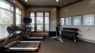 Located at 32 Ardmion Park in Asheville, NC, this fitness center is a thoughtfully designed, single-floor workout facility offering a comprehensive range of exercise equipment and amenities. The space is finished with durable rubber flooring throughout, providing both safety and functionality for a variety of workout routines.

The cardio zone features multiple treadmills and stationary bikes positioned along large, double-hung windows fitted with horizontal blinds, allowing generous natural light to fill the room while providing views of the surrounding outdoor area. Wall-mounted flat-screen televisions accompany the cardio stations for entertainment during workouts.

On the strength training side, the facility includes a full rack of dumbbells organized on a tiered rack, a multi-station cable machine with various attachment options, and a dedicated free weights bench area. Workout reference charts are displayed on the walls, offering exercise guidance for users.

Large wall mirrors span key sections of the room, visually expanding the space and allowing for proper form checks during training. Exposed wooden beams and copper-toned ductwork along the ceiling lend an architectural character to the interior, complementing the warm, neutral-toned walls. A dual water fountain station is conveniently positioned near the strength training area. Recessed lighting provides consistent illumination throughout the facility.