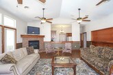This single-floor home offers a cohesive and welcoming interior defined by rich hardwood flooring, warm wood trim, and thoughtful detailing throughout. The main living area is a standout space, anchored by a classic brick fireplace with a wood mantle and flanked by large windows and French doors that flood the room with natural light and provide access to an outdoor patio. A ceiling fan overhead and a wall-mounted television above the fireplace add both comfort and functionality. The space flows naturally into a secondary sitting area, which features vaulted ceilings, additional French doors with transom windows, and half-wall wood paneling that adds a traditional charm. An open pass-through connects this area to the kitchen, making the layout ideal for entertaining. The kitchen is well-equipped, showcasing rich dark cabinetry, dark tile flooring, dark countertops, and a full suite of appliances including a range, microwave, and refrigerator. A chandelier adds an elegant touch overhead. Throughout the home, consistent use of warm wood tones, decorative rugs, and ceiling fans creates a cohesive, comfortable ambiance that is both inviting and functional.