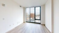 Located on the 12th floor at 801 N Orange St in downtown Wilmington, Delaware, this contemporary 1-bedroom, 1-bathroom residence offers a thoughtfully designed open layout with modern finishes throughout. Light wood-tone flooring flows seamlessly across the entire space, complementing the clean white walls and creating a bright, cohesive aesthetic.

The combined kitchen and living area is anchored by a sleek, well-appointed kitchen featuring flat-front gray cabinetry, white quartz countertops, stainless steel appliances — including a range, microwave, dishwasher, and refrigerator — and a peninsula with an integrated sink, ideal for both meal prep and casual dining.

The generously sized primary bedroom is bathed in natural light courtesy of expansive floor-to-ceiling windows framed in black, offering striking panoramic views of the Wilmington skyline and beyond. The same dramatic window treatment carries through the living area, further amplifying the sense of openness and connecting the interior to the urban landscape outside.

The bathroom is finished with large-format marble-look tile surrounding a soaking tub and shower combination, complemented by a wood-toned vanity and gray floor tile, striking a balance between elegance and function. A dedicated closet provides practical storage. The overall ambiance is modern, airy, and sophisticated.