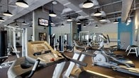 This fitness center is designed to support a comprehensive workout experience within a thoughtfully designed, industrial-style interior. The space features exposed ductwork and structural elements overhead, painted in neutral tones that complement the open, warehouse-inspired aesthetic. A grid of pendant light fixtures with black shades provides ample, evenly distributed illumination throughout the floor.

The cardio section is prominently equipped with a row of treadmills, elliptical trainers, and stationary bikes, all arranged to maximize use of the space. Mounted flat-screen televisions are positioned for easy viewing during workouts, adding a modern convenience to the environment. Floor-to-ceiling mirrors line the walls, visually expanding the space and allowing users to monitor their form during exercise.

Beyond the cardio area, the strength training zone offers a variety of cable machines and weight equipment, catering to a full range of fitness goals. The flooring is dark and durable, appropriate for high-traffic athletic use. Structural columns with bold accent colors add a design element that breaks up the open floor plan. Overall, the space combines functional equipment density with an energetic, modern ambiance suited for serious fitness activity.