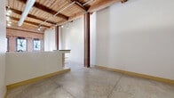 This character-rich urban loft showcases a thoughtful conversion of a historic building, blending industrial heritage with modern livability across a single floor. The open-concept living, dining, and kitchen area serves as the heart of the space, where exposed red brick walls, original heavy timber beam ceilings, and polished concrete floors create a striking industrial aesthetic. Large windows along the brick wall flood the living area with abundant natural light, while a ceiling fan adds practical comfort. The kitchen is outfitted with light maple cabinetry, dark granite countertops, stainless steel appliances including a range and over-the-range microwave, and a peninsula that defines the cooking zone. Hallways connect the living area to two bedrooms, each featuring the same concrete flooring and wood beam ceilings, with one room incorporating a built-in platform element. The two bathrooms include a full bath with a distinctive curved walk-in shower flanked by glass block panels, and a second bath featuring a double-sink maple vanity with a dark countertop and matte black fixtures. Additional utility and laundry rooms, along with two closets, provide practical storage. Exposed structural columns and industrial conduit throughout reinforce the loft's authentic, converted character.