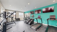 Located at 38 Westgate Dr. in Dover, NH, this fitness center offers a well-organized, fully equipped exercise space designed to support a comprehensive workout routine. The room features a clean, modern aesthetic highlighted by a bold teal accent wall that adds a vibrant pop of color against the neutral gray walls and light ceiling. Recessed lighting throughout ensures consistent, even illumination, while multiple large windows along two walls flood the space with natural light and provide views of the surrounding property.

The facility is outfitted with a diverse selection of equipment to accommodate both cardio and strength training. Cardio offerings include multiple TRUE brand treadmills, stair climbers, and elliptical machines. For strength training, the room provides a multi-functional cable machine, a Smith machine or cable-and-pulley system with an adjustable bench, and a well-stocked dumbbell rack with a full range of free weights. Two flat-screen TVs are mounted on the teal accent wall, allowing residents to stay entertained during workouts.

The durable rubberized flooring offers safety and comfort underfoot. A mini-split HVAC unit ensures year-round climate control, and a dedicated entryway with glass-panel doors provides convenient access. The space is thoughtfully arranged to maximize usability and flow throughout the room.