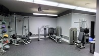 This single-floor fitness space is designed to support a comprehensive strength training experience. The room features light gray walls accented by a bold dark gray upper band near the ceiling, creating a clean, modern aesthetic. Durable rubber flooring covers the entire area, providing both safety and comfort during workouts. The space is furnished with a selection of professional-grade equipment, including a multi-function cable machine, a chest press station, a leg press or seated strength machine, and an adjustable weight bench, offering a well-rounded range of upper and lower body exercises. Overhead fluorescent lighting ensures the entire space is brightly and evenly illuminated, while a ceiling fan promotes air circulation during intense sessions. Exposed metal ceiling tracks add an industrial touch to the overall design. Wall-mounted storage or bar fixtures along the perimeter make efficient use of the available space. The layout is thoughtfully arranged to allow clear movement between machines, supporting a smooth and practical workout flow. Overall, this fitness space combines functional design with quality equipment to deliver a capable and comfortable training environment.