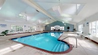 This indoor pool facility is designed to provide a comfortable and functional aquatic experience year-round. The centerpiece is a generously sized swimming pool with a classic brick-tiled border, accessible via wide entry steps fitted with safety handrails. Adjacent to the main pool is a circular hot tub, offering a relaxing complement to the swimming area. The soaring vaulted ceiling, accented by exposed white beams and strategically placed skylights, fills the space with abundant natural light, creating an airy and open atmosphere. Large windows line the perimeter walls, framing views of the surrounding greenery and reinforcing the bright, welcoming ambiance. The expansive pool deck is finished in light-toned concrete, providing ample space for movement and relaxation. A row of poolside lounge chairs and additional seating areas invite guests to unwind. A dedicated area along one wall includes a shelving unit stocked with water fitness equipment such as foam noodles, kickboards, and dumbbells. The facility also features multiple changing rooms and restroom facilities, as well as a ceiling fan for added ventilation. Safety equipment, including a life ring station, is prominently displayed, and pool rules signage is posted throughout for the convenience of all guests.