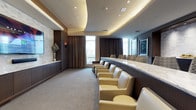 This private screening room is a study in refined sophistication, thoughtfully designed to deliver an upscale entertainment experience. The space is anchored by a striking tray ceiling with warm cove lighting and recessed downlights, creating a layered, atmospheric glow that sets the mood while maintaining a polished aesthetic. A sleek marble-topped bar counter runs along one side of the room, offering a generous surface ideal for hosting guests in style. Plush cream leather armchairs are arranged in a row along the viewing area, providing comfortable, lounge-style seating that invites relaxation. The walls are finished with rich dark wood paneling and complemented by textured stone accent panels, adding depth and warmth to the interior. Neutral-toned carpeting runs underfoot, softening the space acoustically and visually. Floor-to-ceiling windows dressed with floor-length chocolate brown drapes allow for natural light when desired while offering complete blackout capability for optimal viewing conditions. Framed artwork adorns the walls, adding a curated, gallery-like touch. The overall ambiance is one of quiet luxury — intimate yet spacious, and purpose-built for an exceptional private cinema experience.