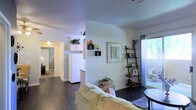 This single-floor residence offers a warm and functional living environment with a thoughtfully designed open-concept layout. The living area features rich dark hardwood floors that contrast elegantly with bright white walls, creating a clean and inviting ambiance. A plush sofa anchors the sitting space, complemented by a decorative leaning ladder shelf that provides both style and storage. Framed wall art and tasteful accents add personality throughout the room.

The living area flows seamlessly into the kitchen, which is equipped with white cabinetry extending to the upper wall and a full-size white refrigerator. The raised upper cabinetry provides generous storage, and the kitchen's open sightlines maintain a sense of connectivity with the rest of the home.

Natural light fills the space through a large sliding glass door that leads to an outdoor area, offering views of lush greenery beyond. A ceiling fan in the adjoining hallway area enhances comfort and air circulation. The hallway connects to additional rooms, including what appears to be a bedroom, maintaining a practical and easy-to-navigate floor plan. Overall, the space balances everyday functionality with a comfortable, homey aesthetic.