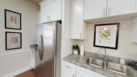 This single-floor clubhouse is designed to serve as a welcoming community hub, combining practical amenities with a clean, modern aesthetic. The kitchen area is well-equipped with crisp white shaker-style cabinetry extending floor to ceiling, providing generous storage capacity. Granite-style countertops offer ample workspace and are complemented by a stainless steel sink, a side-by-side refrigerator with an in-door ice and water dispenser, and a dedicated coffee station. The continuity of white cabinetry and consistent hardware throughout creates a cohesive, polished look.

Adjacent to the kitchen, a large central island with matching countertops and white cabinetry serves as a natural gathering point. The open common area features warm hardwood-style flooring, neutral wall tones, and recessed panel wainscoting that adds architectural detail. Pendant lighting and wall sconces contribute to a well-lit, inviting atmosphere. Upholstered seating arrangements provide comfortable lounge areas, while a dining table with upholstered chairs offers a more formal setting near a fireplace. A wall-mounted television adds entertainment value. Multiple windows along the perimeter allow natural light and views into the adjacent fitness area, enhancing the sense of openness throughout the space.