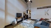 This single-floor space showcases a striking industrial-modern aesthetic defined by dramatically high ceilings that lend an expansive, airy quality to the interior. Exposed ductwork and bold black ceiling beams contrast beautifully against crisp white walls, reinforcing the loft-inspired character of the space. A ceiling fan with wide blades complements the vertical scale of the room while enhancing air circulation throughout.

The living area is anchored by polished concrete floors and features a large bank of windows fitted with wooden horizontal blinds and sheer roller shades, allowing for flexible control of the abundant natural light that floods the space. Views through the oversized windows reveal an urban streetscape, connecting the interior to its city surroundings. An infrared wall-mounted heater is visible near the window wall, adding a functional yet unobtrusive element.

The living space itself is thoughtfully arranged, with a rustic wood media console, a clean white coffee table, and a patterned area rug grounding the seating area. A corridor leads further into the unit, hinting at additional rooms beyond. The overall ambiance is modern and urban, balancing raw industrial details with comfortable, livable design.