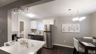 This single-floor space showcases a thoughtfully designed interior with a clean, modern aesthetic. The kitchen serves as a central highlight, featuring a combination of white upper cabinetry and dark lower cabinets that creates a striking two-tone contrast. Stainless steel appliances, including a full-size refrigerator and dishwasher, lend a sleek, contemporary feel. A light-toned stone countertop extends along the island, offering additional prep and gathering space. The sink is positioned beneath a window fitted with white blinds, allowing natural light to filter into the space. Dark hardwood flooring runs throughout, grounding the neutral gray walls and providing a rich, cohesive look. Track lighting and pendant fixtures ensure the space is well-lit and functional. A dedicated laundry area is visible adjacent to the kitchen, adding practical convenience. Decorative wall art with a muted, natural motif complements the overall neutral color palette. The open layout between the kitchen and adjoining dining area promotes an easy flow, making the space feel connected and versatile. Overall, the space presents a polished, move-in-ready environment with a balance of form and function.