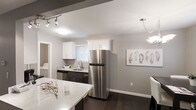This single-floor space showcases a thoughtfully designed interior with a clean, modern aesthetic. The kitchen serves as a central highlight, featuring a combination of white upper cabinetry and dark lower cabinets that creates a striking two-tone contrast. Stainless steel appliances, including a full-size refrigerator and dishwasher, lend a sleek, contemporary feel. A light-toned stone countertop extends along the island, offering additional prep and gathering space. The sink is positioned beneath a window fitted with white blinds, allowing natural light to filter into the space. Dark hardwood flooring runs throughout, grounding the neutral gray walls and providing a rich, cohesive look. Track lighting and pendant fixtures ensure the space is well-lit and functional. A dedicated laundry area is visible adjacent to the kitchen, adding practical convenience. Decorative wall art with a muted, natural motif complements the overall neutral color palette. The open layout between the kitchen and adjoining dining area promotes an easy flow, making the space feel connected and versatile. Overall, the space presents a polished, move-in-ready environment with a balance of form and function.