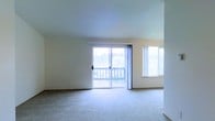 This single-floor space features a spacious, open living area finished with wall-to-wall carpet flooring in a neutral tone, contributing to a clean and understated aesthetic. The room is bathed in natural light, courtesy of a large sliding glass door that opens directly onto an outdoor balcony, offering views of the surrounding landscape including what appears to be lush greenery in the distance. Adjacent to the sliding door, an additional window further enhances the brightness of the space, ensuring the interior feels airy and well-lit throughout the day. The walls are finished in a soft, warm white tone that complements the light carpeting and reinforces the overall sense of openness. The ceiling is smooth and uniform, adding to the clean, simple design of the room. The sliding glass door provides both a functional connection to the exterior and a visual focal point within the living area. Overall, the space presents a straightforward, comfortable layout well-suited for everyday living, with its abundance of natural light and outdoor access serving as key highlights.