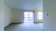 This single-floor space features a spacious, open living area finished with wall-to-wall carpet flooring in a neutral tone, contributing to a clean and understated aesthetic. The room is bathed in natural light, courtesy of a large sliding glass door that opens directly onto an outdoor balcony, offering views of the surrounding landscape including what appears to be lush greenery in the distance. Adjacent to the sliding door, an additional window further enhances the brightness of the space, ensuring the interior feels airy and well-lit throughout the day. The walls are finished in a soft, warm white tone that complements the light carpeting and reinforces the overall sense of openness. The ceiling is smooth and uniform, adding to the clean, simple design of the room. The sliding glass door provides both a functional connection to the exterior and a visual focal point within the living area. Overall, the space presents a straightforward, comfortable layout well-suited for everyday living, with its abundance of natural light and outdoor access serving as key highlights.