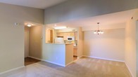 This single-floor residence offers an open-concept layout that seamlessly connects the kitchen and living areas, creating a spacious and functional environment. The kitchen is outfitted with light wood cabinetry, a breakfast bar peninsula, and includes essential appliances such as a refrigerator, microwave, and range. A fluorescent ceiling fixture provides bright, even task lighting over the workspace. The kitchen flows naturally into the adjoining dining and living areas, which feature soft, neutral-toned carpeting underfoot and warm beige walls that lend the space a calm, welcoming ambiance. A decorative chandelier serves as a charming focal point in the dining area, adding a touch of elegance to the open space. The high ceilings amplify the sense of openness and airiness throughout. The clean, understated interior design allows the architectural details — including the open pass-through kitchen design and the smooth wall finishes — to stand out. The overall aesthetic is tidy and versatile, offering a blank canvas well-suited to a variety of décor styles. The layout promotes easy movement and visual connectivity between living spaces, making the home feel both practical and comfortable.