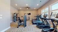 Located at 321 E Park Rd in Indian Trail, North Carolina, this well-equipped fitness center offers a comprehensive workout environment designed to support a wide range of exercise routines. The single-floor space features neutral-toned walls, patterned carpet tiles in earthy tones, and recessed lighting throughout, creating a clean and functional atmosphere.

The cardio section is anchored by a row of True-brand treadmills and elliptical machines, positioned along large, white-framed windows that allow abundant natural light to fill the space and provide views of the surrounding community. A wall-mounted television nearby keeps users entertained during longer sessions.

The strength training area is equally well-appointed, offering a multi-functional cable machine, a leg extension/curl machine, a power rack with a flat bench and weighted barbells, and a dedicated dumbbell rack with a utility bench. Additional wall-mounted televisions are positioned throughout to ensure visibility from multiple workout zones.

Amenities include a water fountain, a sanitizing station for equipment cleaning, and a conveniently accessible restroom. An overhead ceiling fan provides supplemental air circulation. The layout is thoughtfully organized to allow ample space between equipment, supporting safe and comfortable use for multiple occupants simultaneously.