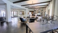 This expansive single-floor space presents a beautifully designed communal lounge area that seamlessly blends transitional and contemporary design elements. The ceiling is a standout architectural feature, adorned with dark-stained exposed wood beams that contrast elegantly against the crisp white ceiling, adding warmth and character to the space. Two distinctive chandeliers — one branch-inspired and one more traditional — serve as focal points overhead, contributing to the refined ambiance.

The flooring transitions between large-format tile and rich dark hardwood, subtly defining different zones within the open layout. Multiple seating arrangements are thoughtfully curated, featuring a mix of plush sofas, accent chairs, and occasional tables that invite relaxed gathering and conversation. Built-in shelving units along one wall display tasteful décor and frame a wall-mounted television, adding both function and visual interest.

Natural light flows generously through multiple windows and glass French doors, connecting the interior to the outdoors. The overall atmosphere is welcoming and polished, reflecting a well-appointed communal environment designed for comfort and social interaction.