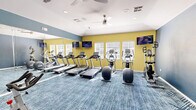 Located at 12055 Sabo Rd in Houston, TX, this fitness center offers a dedicated exercise space designed to support a well-rounded workout routine. The room is finished with a vibrant two-tone color scheme, pairing cool steel blue with bold yellow accent walls, creating an energizing atmosphere for residents and guests alike.

The space is outfitted with a varied selection of cardio equipment, including multiple treadmills, elliptical machines, stationary cycling bikes, and an upright climbing machine. Floor-to-ceiling mirrors along one wall visually expand the room while allowing users to monitor their form during exercise. Patterned blue carpet tiles cover the floor throughout, adding both comfort and a modern aesthetic.

Natural light filters in through windows fitted with white blinds on the yellow accent wall, while dual ceiling fans with integrated lighting ensure consistent air circulation and comfort. Wall-mounted flat-screen televisions are positioned for easy viewing from the cardio equipment. Recessed lighting and HVAC vents in the ceiling contribute to a clean, well-maintained environment.

A small accessory station near the cycling area provides towels and amenities for added convenience. The overall design is practical yet polished, offering a comfortable and fully functional fitness environment.