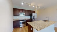 Located along River Shore Drive in Louisville, Kentucky, this single-floor residence offers a well-appointed, open-concept layout designed for functional everyday living. The kitchen serves as the central feature of the space, showcasing rich dark cabinetry, speckled granite countertops, and a full suite of stainless steel appliances, including a range, built-in microwave, and a French door refrigerator with ice and water dispenser. A peninsula with an integrated sink and additional granite surface extends into the open living area, providing a natural division between cooking and living spaces while maintaining visual continuity. Track lighting runs throughout the main living area and kitchen, providing adjustable, well-distributed illumination. Light hardwood-style flooring spans the main living areas, complementing the neutral wall tones and contributing to the bright, airy feel of the space. A dedicated bedroom features a large window that admits generous natural light and offers outdoor views of surrounding greenery, alongside a walk-in closet with wire shelving systems for organized storage. The bathroom includes a granite-topped vanity with dark cabinetry, a tub-shower combination, and hardwood-style flooring. A separate laundry room accommodates a full-size washer and dryer with overhead wire shelving for added utility.