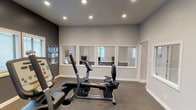 This single-floor community clubhouse is thoughtfully designed to offer both comfort and functionality across its well-appointed spaces. The main lounge area makes a strong first impression with soaring ceilings, an elegant chandelier, and large dark-framed French doors that flood the space with natural light. Neutral-toned furnishings, including a plush cream sofa accented with soft teal and gold pillows, create a warm and inviting atmosphere. A striking floor-to-ceiling stone fireplace with a rustic wooden mantel serves as a centerpiece in the adjacent seating area, complemented by wingback chairs and refined décor. French doors flanking the fireplace open to an outdoor area, enhancing the connection between interior and exterior spaces. The lounge flows seamlessly into a casual dining nook with a wooden table and upholstered chairs, ideal for social gatherings. The fitness center is equipped with cardio machines, including a recumbent bike and treadmill, and features carpeted flooring, recessed lighting, and interior windows that provide visibility into neighboring spaces. Throughout the clubhouse, a cohesive design palette of soft whites, warm grays, and natural stone elements creates a polished yet approachable ambiance suited for community living.