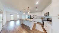 Located at 7785 Griffon Ln in Vero Beach, Florida, this well-appointed two-bedroom, two-bathroom single-floor home offers a clean, modern aesthetic throughout. The kitchen serves as a standout centerpiece, boasting white shaker cabinetry, granite countertops, a large central island with seating, stainless steel appliances, and elegant pendant lighting — all set atop warm hardwood-style flooring that flows seamlessly into the main living area. The living space is bright and airy, enhanced by a ceiling fan, large windows with slatted blinds, and a glass door that invites natural light and views of the surrounding lush greenery. Both bedrooms are carpeted for comfort and feature windows overlooking the verdant outdoor landscape, with ceiling fans adding to the relaxed Florida feel. The primary bedroom connects directly to a dedicated en-suite bathroom, which includes a dual-sink vanity with granite countertops, white cabinetry, a glass-enclosed walk-in shower, and hardwood-style flooring. The second full bathroom is equally well-finished, maintaining the same crisp, white design language. Hallways are clean and open, with raised-panel doors adding a subtle architectural detail. The overall ambiance is light, fresh, and move-in ready.