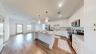 Located at 7785 Griffon Ln in Vero Beach, Florida, this well-appointed two-bedroom, two-bathroom single-floor home offers a clean, modern aesthetic throughout. The kitchen serves as a standout centerpiece, boasting white shaker cabinetry, granite countertops, a large central island with seating, stainless steel appliances, and elegant pendant lighting — all set atop warm hardwood-style flooring that flows seamlessly into the main living area. The living space is bright and airy, enhanced by a ceiling fan, large windows with slatted blinds, and a glass door that invites natural light and views of the surrounding lush greenery. Both bedrooms are carpeted for comfort and feature windows overlooking the verdant outdoor landscape, with ceiling fans adding to the relaxed Florida feel. The primary bedroom connects directly to a dedicated en-suite bathroom, which includes a dual-sink vanity with granite countertops, white cabinetry, a glass-enclosed walk-in shower, and hardwood-style flooring. The second full bathroom is equally well-finished, maintaining the same crisp, white design language. Hallways are clean and open, with raised-panel doors adding a subtle architectural detail. The overall ambiance is light, fresh, and move-in ready.