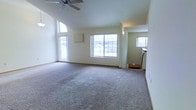 This single-floor home offers a welcoming and open layout designed for comfortable everyday living. The main living area is a standout feature, boasting soaring vaulted ceilings that create an impressive sense of height and volume. A ceiling fan is centrally positioned overhead, providing both functional airflow and a subtle decorative touch. The space is finished with plush wall-to-wall carpeting in a neutral tone, lending warmth and comfort underfoot throughout.

Natural light floods the interior through multiple large windows, including a prominent multi-pane window along the main wall and a tall vertical window that draws the eye upward, further emphasizing the dramatic ceiling height. These windows frame views of the surrounding neighborhood, keeping the space feeling connected to the outdoors. Soft, neutral wall tones complement the light-filled atmosphere, creating a clean and versatile backdrop.

A wall-mounted unit and a sconce light fixture add functional and aesthetic detail to the living area. A glimpse into an adjoining space reveals additional windows and what appears to be a kitchen or utility area, suggesting a practical and well-connected floor plan. Overall, this home exudes a bright, airy ambiance with classic residential design elements throughout.