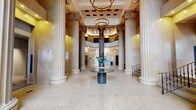 Located at 55 Elm Street in Hartford, Connecticut, this magnificent hallway serves as the grand lobby of a distinguished neoclassical building. The space commands immediate attention with its impressive collection of towering Ionic and fluted columns — some crafted from richly veined dark marble, others finished in warm limestone — that rise dramatically toward the soaring ceiling above. Underfoot, expansive polished terrazzo and stone tile flooring extends throughout, reflecting the ambient glow of recessed ceiling lights and elegant chandelier fixtures. The warm, neutral tones of the stone-clad walls lend the interior a timeless, stately character. Large multi-pane windows at the main entrance flood the lobby with natural light, further highlighting the architectural grandeur of the space. A sleek circular table with decorative greenery serves as a welcoming focal point near the entry. The lobby provides access to both a stairwell with ornate iron railings and a modern elevator bank, ensuring seamless vertical circulation. Visible signage for the 55 Elm Club hints at the building's active, community-oriented character. The overall ambiance is one of refined civic elegance, blending historic architectural heritage with contemporary functionality.