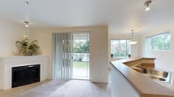 This single-floor residence offers a welcoming and light-filled interior with a thoughtfully designed open-concept layout. The living area is finished with plush wall-to-wall carpeting and neutral warm-toned walls that create a calm, inviting ambiance throughout. A classic white fireplace serves as a focal point in the living room, adding a touch of traditional charm to the space.

Natural light flows generously into the living area through a full-length sliding glass door fitted with vertical blinds, which opens directly onto an outdoor patio overlooking a lush green yard bordered by a white lattice fence. This seamless indoor-outdoor connection enhances the sense of openness and brings the surrounding greenery into view.

Adjacent to the living room, the kitchen area is defined by a raised counter that acts as a natural divider between spaces. A pendant light fixture hangs above, adding warmth and definition to the kitchen zone. A window with a view of outdoor foliage further brightens the kitchen, making it an airy and functional space.

Overall, the residence balances comfort and practicality, with consistent finishes, ample natural light, and direct access to outdoor spaces that collectively create a serene and livable environment.