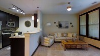 This single-floor residence offers a welcoming, open-concept design that seamlessly connects the kitchen and living areas into one cohesive space. The living room is anchored by a plush, neutral-toned sofa adorned with patterned throw pillows and complemented by a comfortable accent chair draped with a striped throw blanket. A solid wood coffee table with drawer storage adds a rustic warmth to the space, while a soft carpet underfoot enhances the cozy atmosphere. Overhead, a ceiling-mounted light fixture provides ambient illumination, and a floor lamp in the corner adds additional layered lighting. Decorative wall art, including a classic Parisian cityscape print and subtle gear-shaped accents, gives the living space a curated, personalized feel. The adjacent kitchen area features a raised granite-topped breakfast bar with pendant lighting hanging above, offering a casual dining or entertaining option. A large decorative wall clock adds a stylish focal point near the kitchen. The overall color palette is composed of soft beiges, warm grays, and golden yellow accents, creating a harmonious and inviting ambiance throughout this thoughtfully designed single-level home.