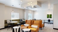 This thoughtfully designed single-floor space functions as a contemporary lounge and coworking area, offering a versatile and welcoming environment. The open layout is anchored by a central seating zone featuring bold amber wingback chairs paired with sleek, geometric side tables, creating an inviting focal point beneath a striking cluster pendant light fixture with globe shades. A built-in green banquette lines one wall, accompanied by round white bistro tables and black café chairs, providing casual dining or meeting areas. Natural light flows generously through multiple windows fitted with horizontal blinds, offering views of the surrounding area and enhancing the bright, airy atmosphere. The neutral palette of soft whites, warm grays, and beige carpeting throughout serves as a clean backdrop for the carefully chosen furnishings. A dedicated workspace area along one wall features long wooden desks and task chairs, ideal for focused work. A private office nook includes a minimalist wood-and-steel desk, pendant lighting, and vibrant moss art panels, adding a natural accent. The space is anchored by a reception counter, with two access doors on either side. Recessed and pendant lighting throughout ensures a well-lit, functional environment suitable for both collaboration and productivity.