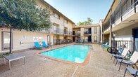 Located in the vibrant Knox-Henderson neighborhood of Dallas, Texas, this courtyard common area at 3516 Matilda St offers a well-appointed outdoor retreat designed for relaxation and community living. The centerpiece of the space is a generously sized swimming pool with clear, blue water, framed by a concrete pool deck finished with terracotta-toned tile edging. Surrounding the pool, a variety of outdoor seating options are thoughtfully arranged, including bright blue lounge chairs, a shaded patio area with a lime-green umbrella and matching accent furniture, and a charming courtyard nook with a seating bench tucked beneath a mature shade tree. A charcoal grill station is conveniently positioned nearby, adding to the outdoor living appeal. The space is enclosed by multi-story residential buildings featuring neutral beige siding, dark-trimmed balconies, and private patios on the ground level separated by black iron railings. Exterior staircases provide access to upper-level units, enhancing the open-air, courtyard-style atmosphere. The mature tree anchoring one corner of the courtyard adds natural character and shade to the overall setting. This outdoor amenity area benefits from open-sky exposure, creating a bright and inviting environment throughout the day.