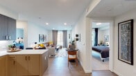 This single-floor residence showcases a thoughtfully designed open-concept layout that seamlessly connects the kitchen, living, and dining areas. The kitchen is equipped with dark wood cabinetry extending to the ceiling, stainless steel appliances including a refrigerator and microwave, and a peninsula with bar seating, providing both functionality and a natural gathering point. Light wood-style flooring runs throughout, complementing the crisp white walls and recessed lighting overhead.

The living area is generously proportioned, anchored by a leather sectional sofa, accent chairs, and a geometric-patterned area rug. Large windows dressed with floor-length drapes allow natural light to fill the space while offering views of the surrounding urban environment. A dedicated workspace with a round desk and chair sits conveniently adjacent to the living area.

The bedroom features a distinctive leather-strap headboard, soft blue bedding, and draping curtains that frame a window, creating a calm, restful atmosphere. A closet with open shelving provides accessible storage nearby.

The bathroom is finished with warm wood-toned cabinetry, a white undermount sink, a large mirror, open shelving for linens, and a ladder-style towel warmer, combining practicality with a clean, modern aesthetic throughout.