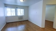 This single-floor space offers a clean, minimalist aesthetic with a straightforward and functional layout. The main room is finished with warm hardwood flooring that extends throughout, adding a natural and inviting quality to the interior. Bright white walls and a smooth ceiling with a central flush-mount light fixture contribute to a bright, open atmosphere. A large window allows natural light to filter in, offering views of mature trees outside, creating a connection to the outdoors. Climate comfort is well-addressed, with a wall-mounted air conditioning unit installed beneath the window and a baseboard heating system running along the perimeter of the room, ensuring year-round temperature control. The room's clean lines and neutral palette provide a versatile canvas, well-suited for a variety of furniture arrangements and personal styling. The space presents a practical and comfortable environment, with essential amenities thoughtfully integrated into the design without compromising the simplicity and openness of the interior.